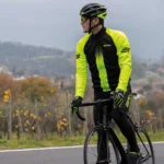 Ciclista in giubbino BRN giallo fluo e nero, mentre pedala su strada, con paesaggio collinare sullo sfondo.
