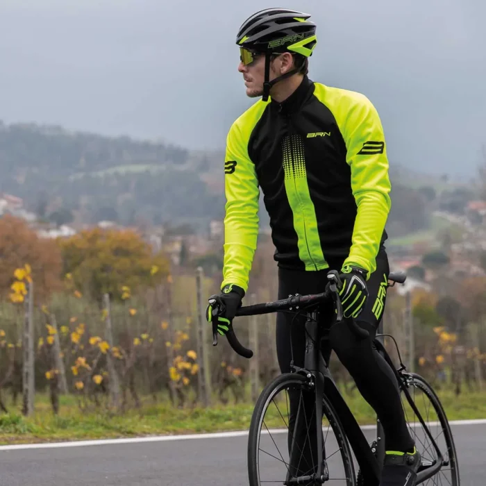 Ciclista in giubbino BRN giallo fluo e nero, mentre pedala su strada, con paesaggio collinare sullo sfondo.