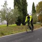 Ciclista in giubbino BRN giallo fluo e nero, che pedala su strada, con alberi di ulivo ai lati