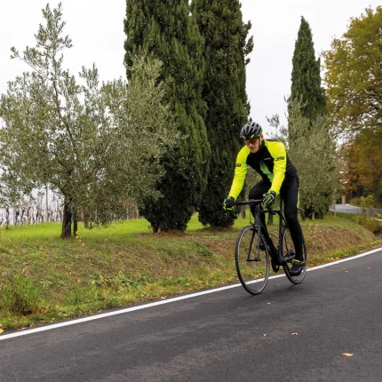 Ciclista in giubbino BRN giallo fluo e nero, che pedala su strada, con alberi di ulivo ai lati