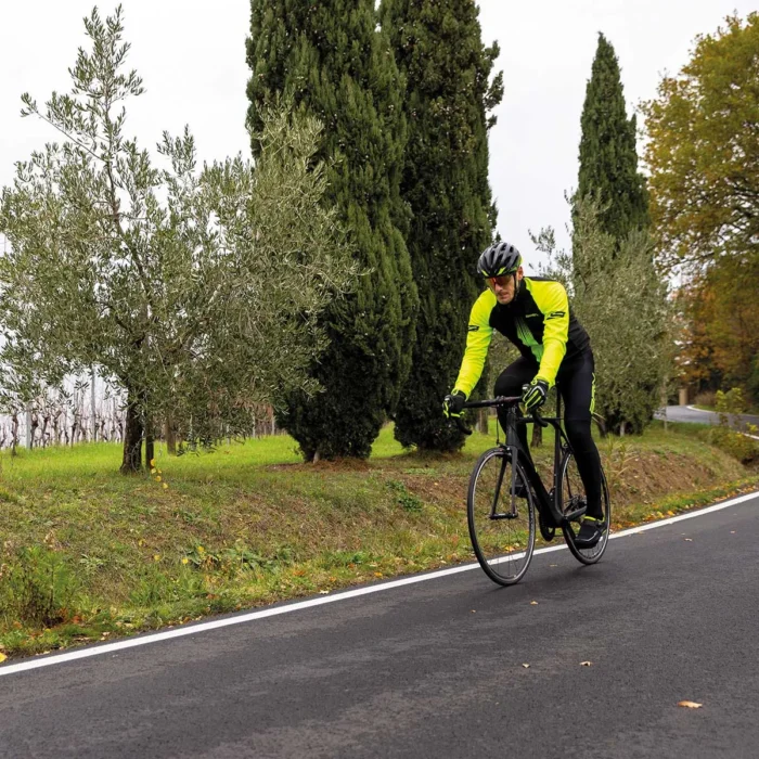 Ciclista in giubbino BRN giallo fluo e nero, che pedala su strada, con alberi di ulivo ai lati
