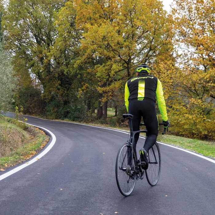 Ciclista di spalle in giubbino BRN giallo fluo mentre affronta una curva su una strada circondata da alberi autunnali.