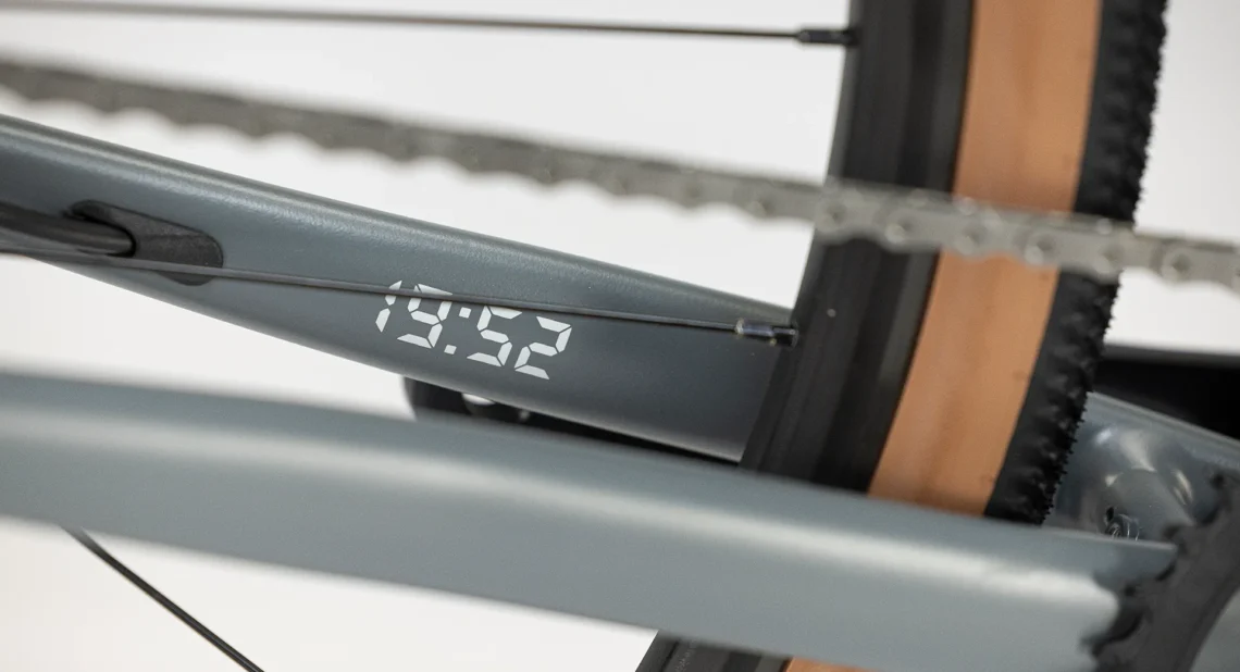 Dettaglio del telaio di una bicicletta grigia, con un display digitale che mostra l'orario '19:52', evidenziando l'integrazione di tecnologia moderna nel design.