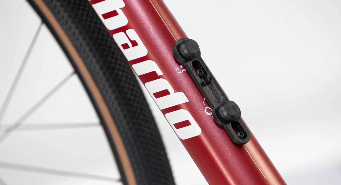 Dettaglio del telaio di una bicicletta rossa, evidenziando il supporto per accessori e il logo del marchio, con una finitura liscia e moderna.