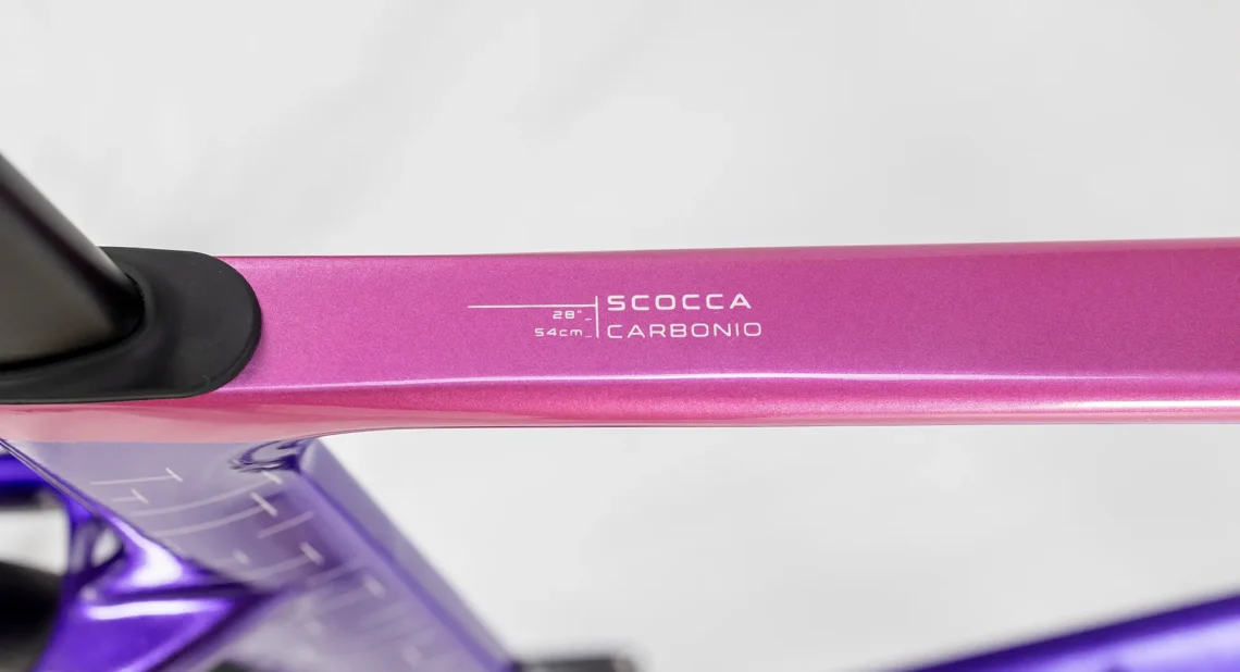 Dettaglio del tubo orizzontale della bicicletta Lombardo, mostrando il modello 'Scocca Carbonio' con misure e angoli precisati. Il design elegante in rosa e viola mette in risalto la qualità del materiale.