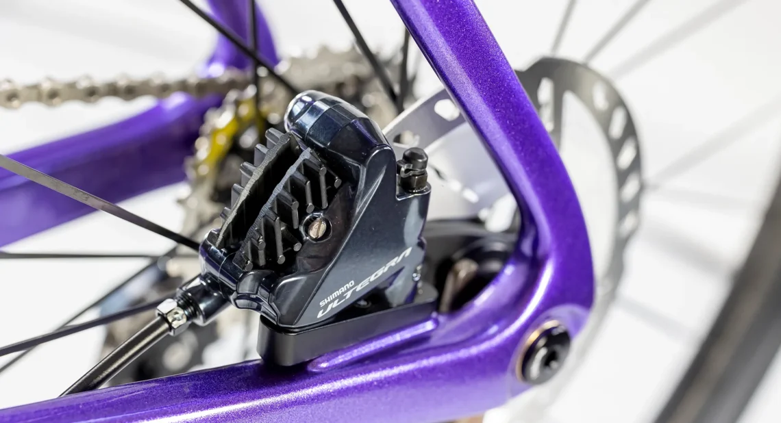 Dettaglio del freno posteriore della bicicletta Lombardo, evidenziando il sistema freno Shimano Ultegra e il design in colore viola del telaio. Mostra la qualità dei componenti e l'attenzione ai dettagli.
